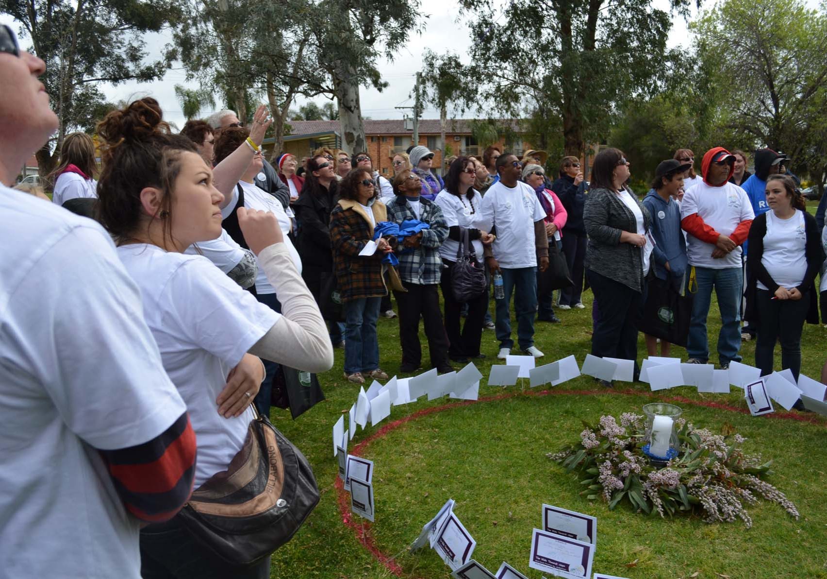 A group of people gathered outdoors, standing around a circle marked with small signs and a candle centerpiece on the grass. Many participants are wearing white shirts.