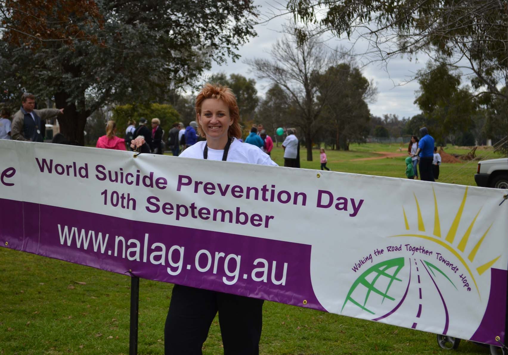 Person holding a banner for World Suicide Prevention Day event on 10th September. Website: www.nalag.org.au. Other people and trees are visible in the background.